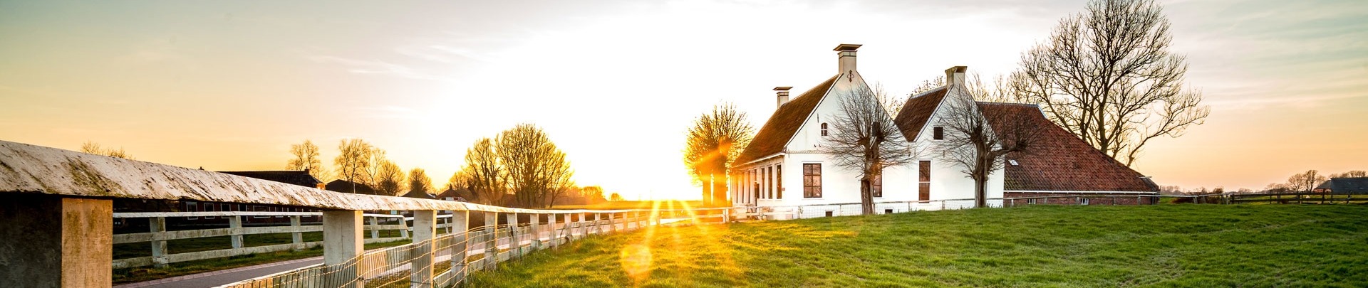 Landweggetje in het Nederlandse landschap met wit hek dat leidt naar een oude boerderij in Nederland tijdens een sfeervolle zonsondergang